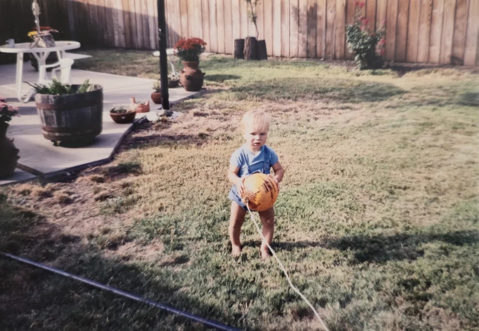 Ken as a toddler holding a ball in a backyard with a wooden fence.