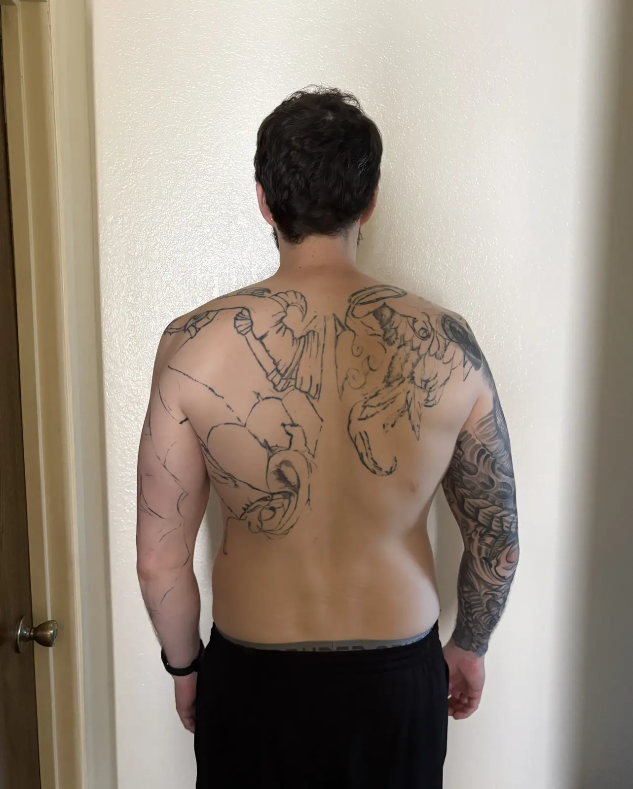 From behind, shirtless, arms down at sides. Same unfinished back tattoo, broader shoulders and more visible back definition, shaded right-arm sleeve.