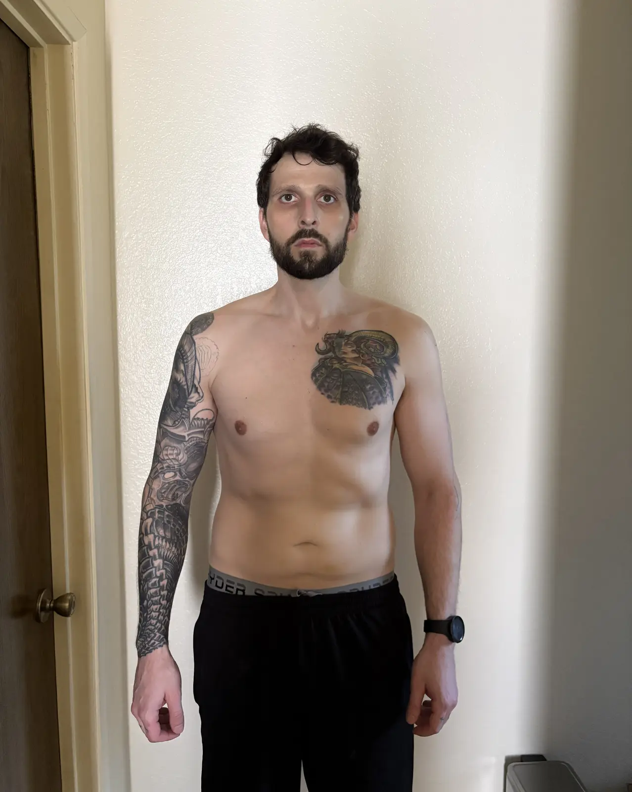 Facing the camera, shirtless, arms at sides. Leaner midsection with more visible definition, trimmed beard, same chest tattoo and sleeve.