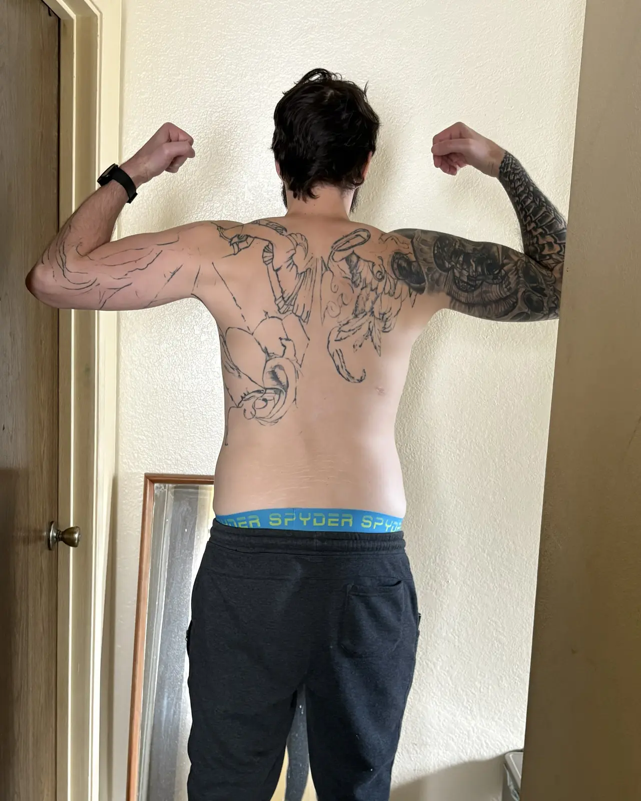 From behind, shirtless, arms raised in a double-biceps flex. Large unfinished line-work tattoo across the upper back, shaded right-arm sleeve.