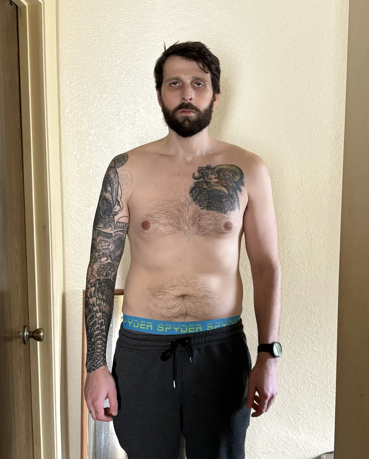 Facing the camera, shirtless, arms at sides. Fuller midsection and chest, full beard, colorful chest tattoo and a full right-arm sleeve.